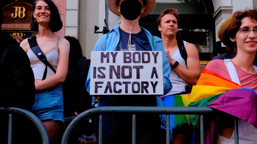Varias personas detrás de una valla en una marcha. Una de ellas sostiene un cartel que dice "My body is not a factory".