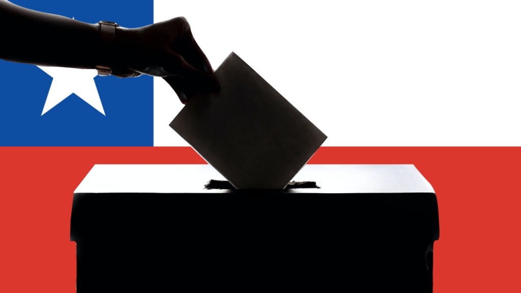 Una mano colocando un sobre en una urna. Detrás la bandera de Chile.