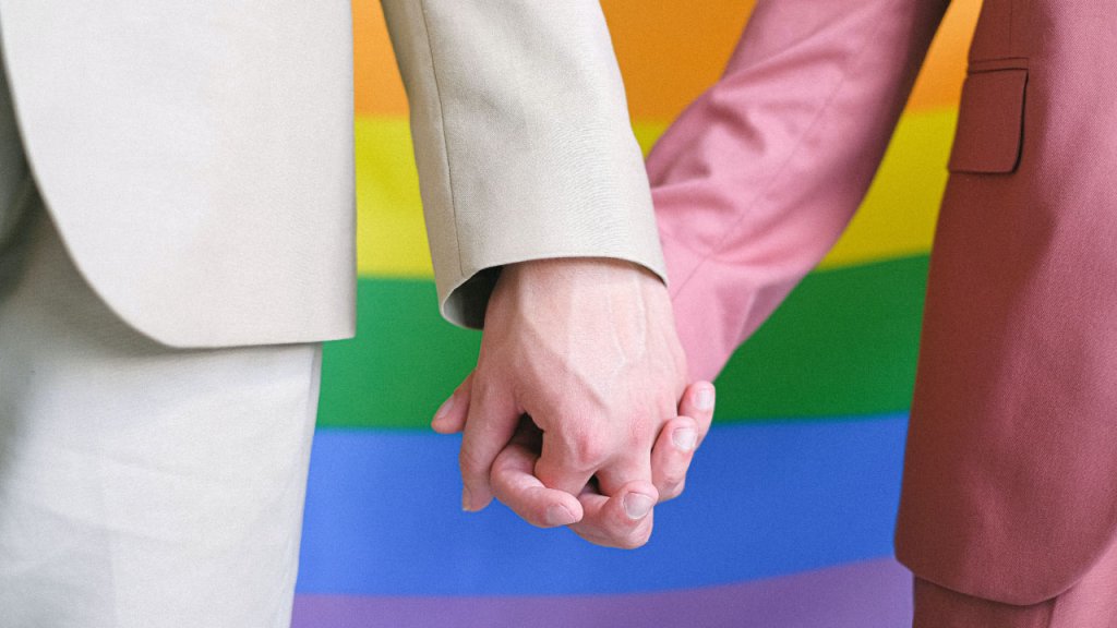 Dos personas tomándose de la mano, una con traje color beige y la otra con traje rosa. De fondo la bandera del Orgullo LGBTIQ+.