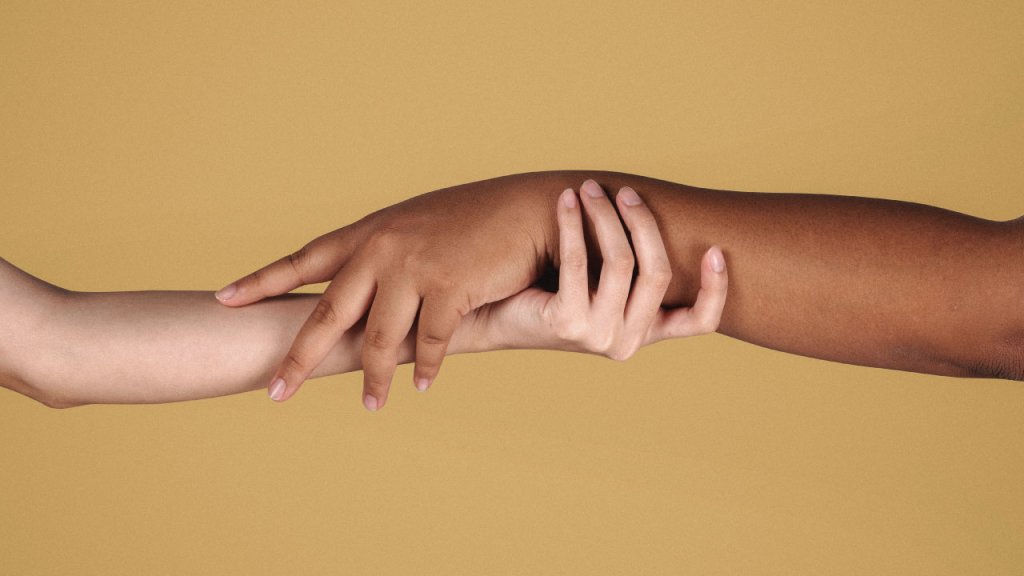 Dos personas tomándose de la mano. De un lado una mano de una persona blanca y del otro lado la mano de una persona marrón.