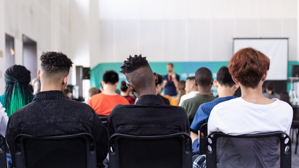 Imagen de un profesor dando una clase a adolescentes. En la imagen se ven las espaldas de les adolescentes y al fondo el profesor.